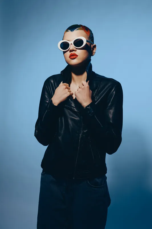 Young woman with colorful hair and white sunglasses wearing black leather jacket posing confidently against blue background, modern style and bold attitude.