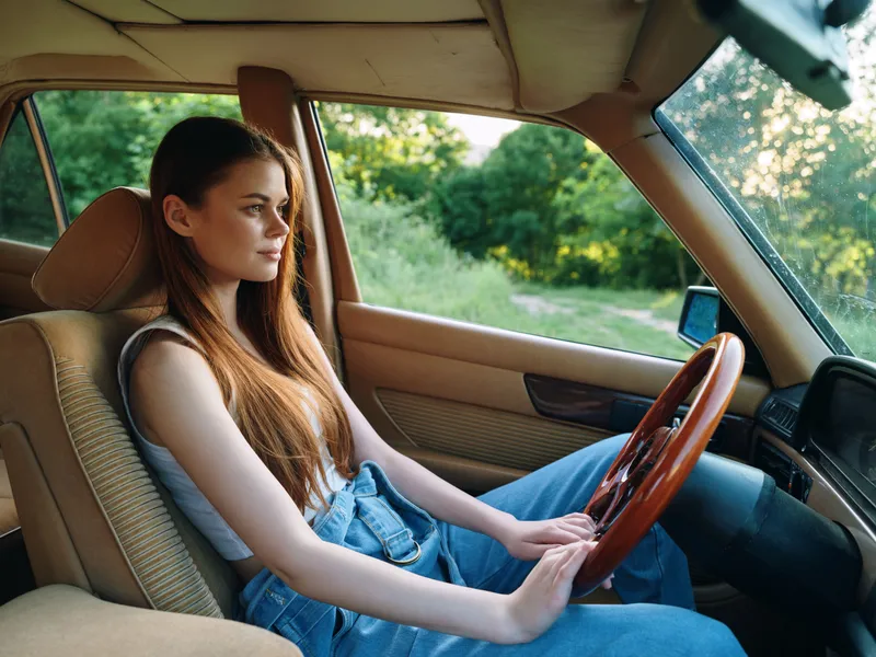 A young woman drives an old car, showcasing a sense of freedom, exploration, and nostalgia in a lush green landscape during a sunny day.