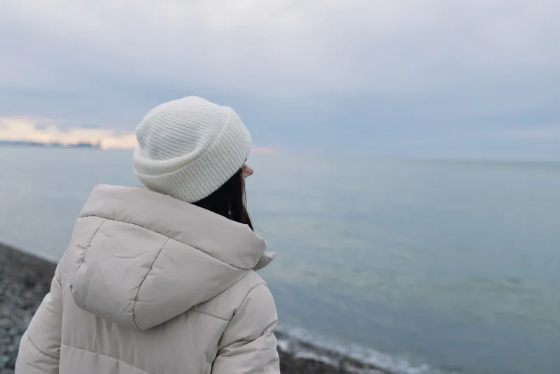 Winter scene of a person in a puffy coat and white beanie gazing at a calm sea along a rocky coastline, soft sky and reflective mood, capturing solitude by the water.