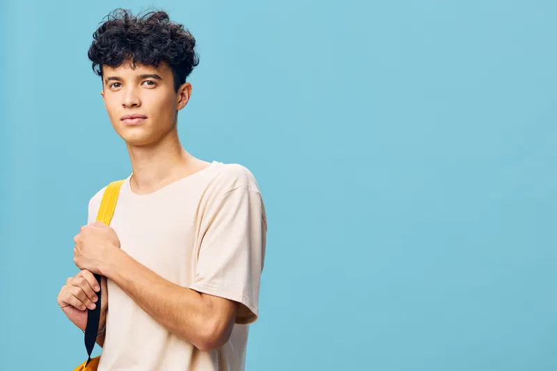 Молодой человек с кудрявыми волосами стоит на фоне светло-голубого цвета, небрежно держа рюкзак