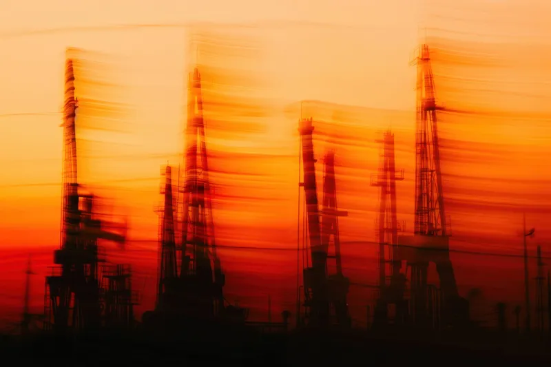 Размытое изображение нефтяных вышек на закате, создающее драматическую атмосферу с яркими оранжевыми и красными оттенками