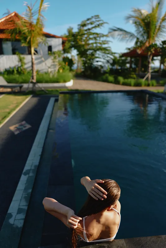 A woman in a swimsuit sits by the pool with her back to the camera, running her hands through wet hair. The tropical setting includes palm trees and a villa in the background.