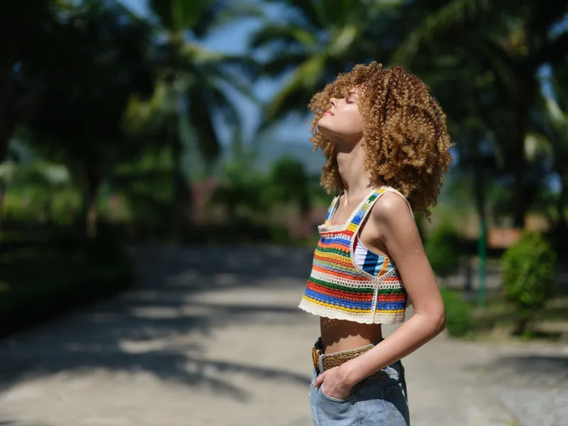 young woman with curly hair wearing colorful crochet top and denim shorts standing outdoors, enjoying warm sunlight with eyes closed, tropical background
