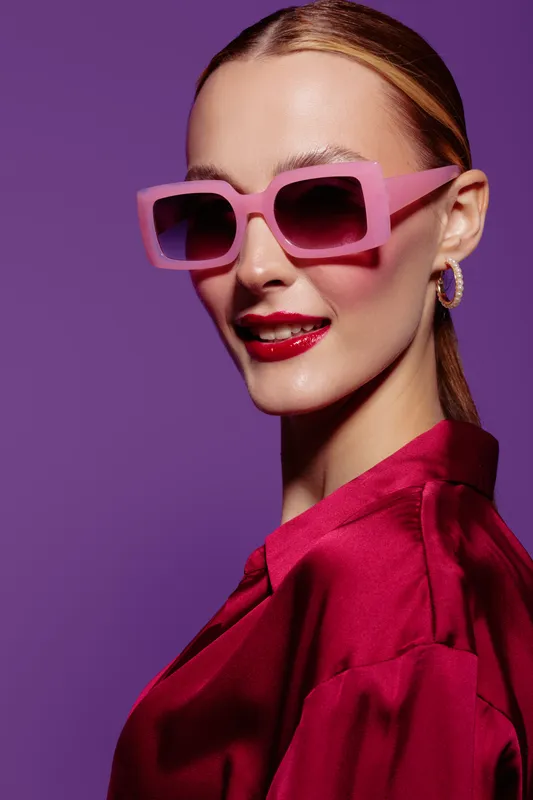 Stylish woman with pink sunglasses and red lipstick smiles confidently against purple background. Fashion concept with trendy accessories and vibrant colors in studio lighting.
