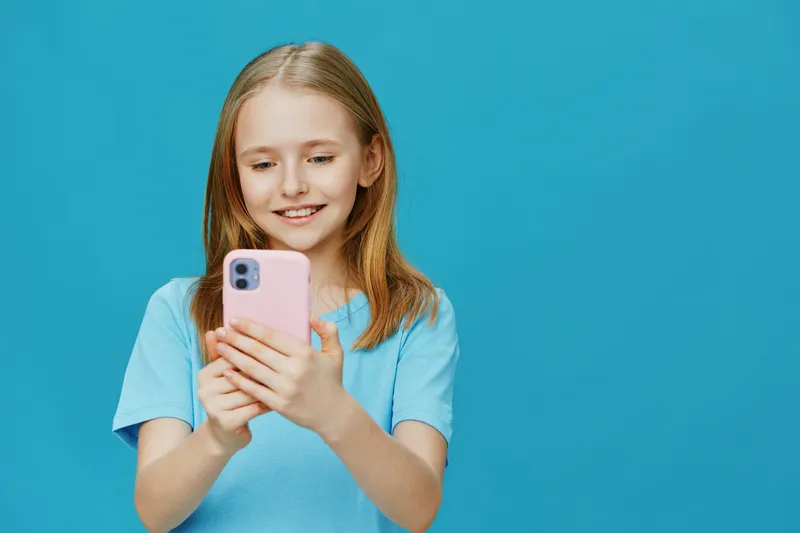 Smiling young girl holding a smartphone in a light blue t shirt against a bright blue background, showcasing joy and technology