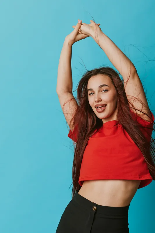Young woman with long hair wearing red crop top and black pants posing with arms raised against blue background. Casual style, smiling and confident female portrait in studio.