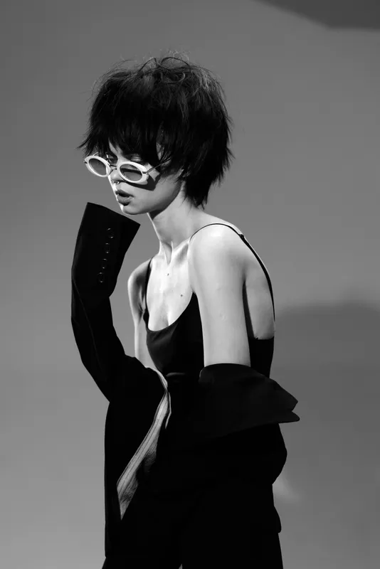 Black and white portrait of a woman with short hair wearing white sunglasses and stylish dark clothing in a minimalist studio setting.
