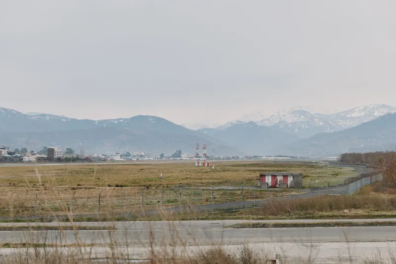 Wide landscape of an empty airfield with grass and small structures in foreground, distant buildings and snow-capped mountains under cloudy sky. Concept of open space and rural environment.