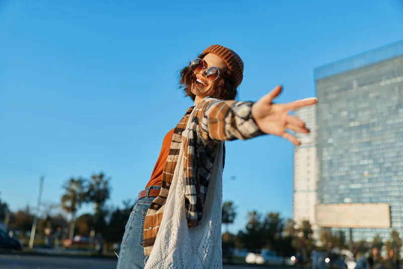 woman smile candid lifestyle golden hour urban joyful portrait of a carefree young woman in beanie and sunglasses, arms outstretched in sunlight glow, emotional storytelling and authenticity vibe.
