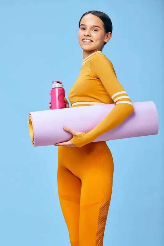 Fitness woman in orange athletic wear holding a yoga mat and water bottle against a bright blue studio backdrop, ready for workout, training, and wellness.