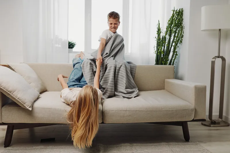 Happy children play on beige sofa in bright living room, boy covered with gray blanket holds smiling girl upside down. Concept of family fun, childhood joy, and indoor activity.