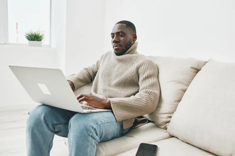 Young African man working on a laptop in a cozy, modern living room setting, showcasing productivity and comfort in a neutral color palette