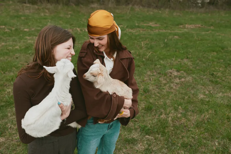 Two women smile while holding goat, goats, and kid in a rural field, enjoying an outdoor lifestyle moment. Warm, natural light highlights friendship, care, and animal companionship in a simple farm