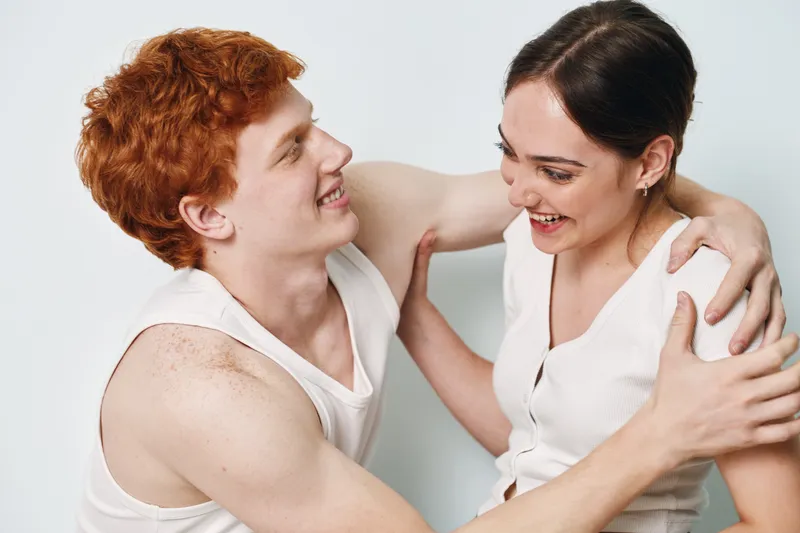Young man and woman smiling and embracing with happy expressions in white tank tops on plain background. Closeup joyful couple laughing and looking at each other with affection and warmth.