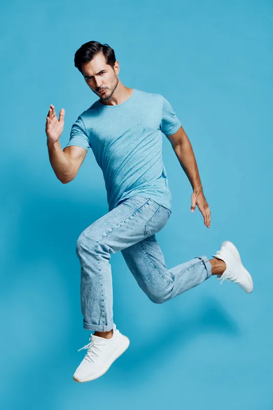 Dynamic athletic man wearing blue t-shirt and jeans running in mid-air with a cheerful expression against solid turquoise background. Active lifestyle concept.