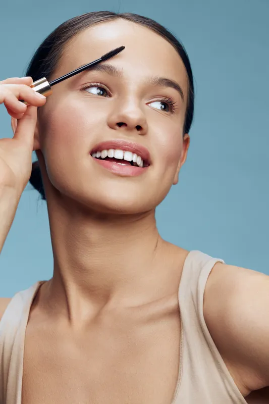 Young woman applying mascara with a joyful expression on a light blue background, showcasing beauty, fashion, and skincare
