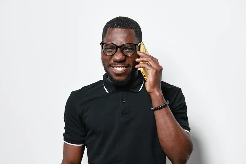 African American man with dark skin wearing glasses and black polo shirt talking on smartphone smiling against white background. Casual communication and technology concept.