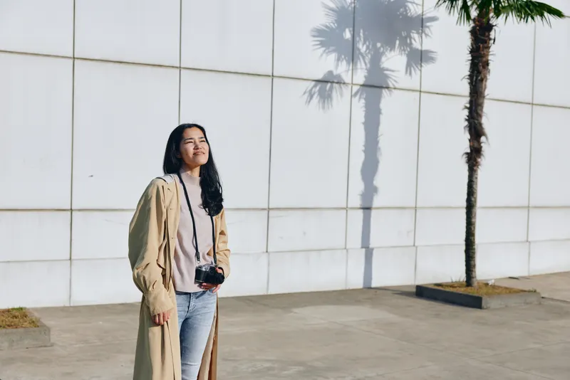 Woman with camera standing beside a white tiled wall under palm shade, casual street style, outdoor photography vibe, warm light, fashion moment in urban setting