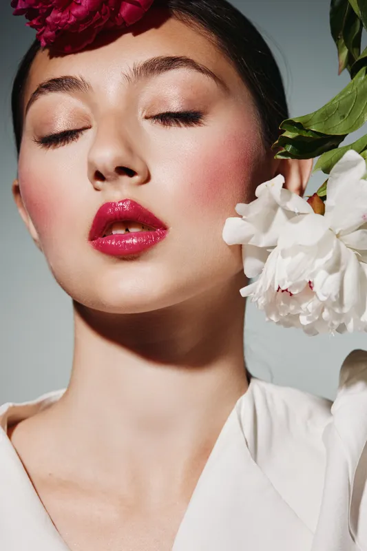 Young woman with glowing skin, bold red lips, and creative floral headpieces. Close-up beauty fashion portrait in natural light. Concept for fashion editorial and cosmetics.