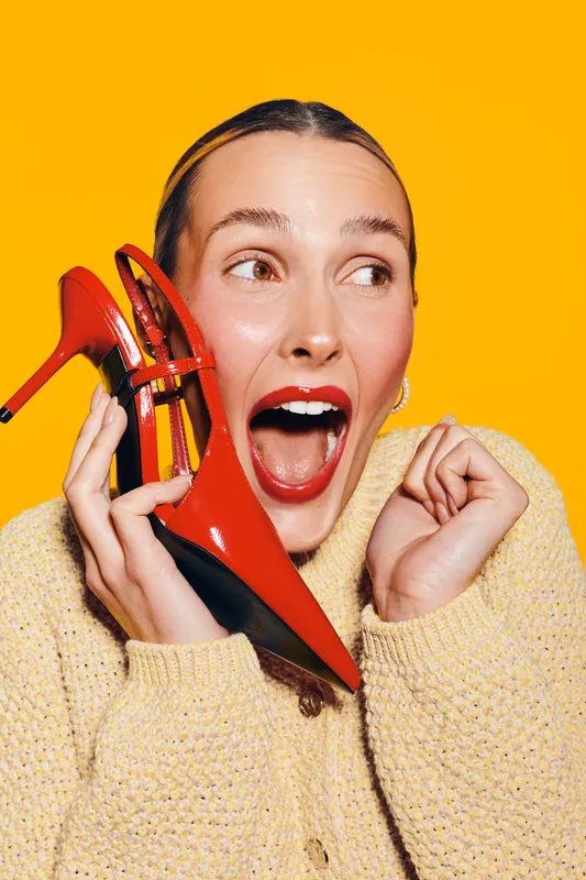 Excited woman in yellow sweater holds red high heel shoe near face, mouth open in surprise. Bright yellow background creates vibrant, joyful mood. Concept for fashion and happiness.