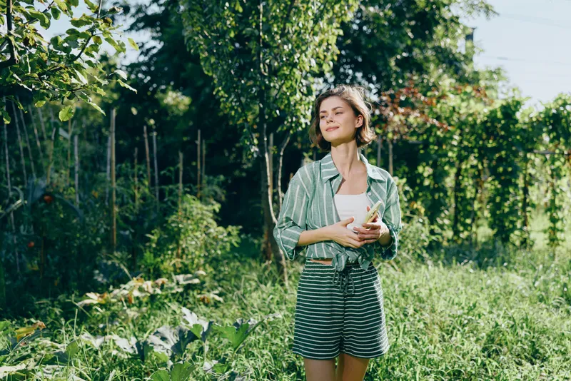 woman stands outdoors amid lush greenery in casual outfit, striped shorts and light jacket, holding a smartphone, sunlit scene, serene mood, natural backdrop, relaxed pose, fashion, leisure
