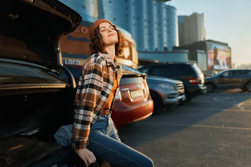 woman car parking trunk sunlight smile urban candid portrait of a young woman leaning on an open car trunk in golden hour glow, authenticity, mindful living and emotional storytelling