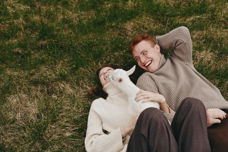 Young couple lying on grass smiling and playing with a white baby goat in a cozy autumn setting, wearing knitted sweaters and enjoying a relaxed lifestyle moment outdoors.