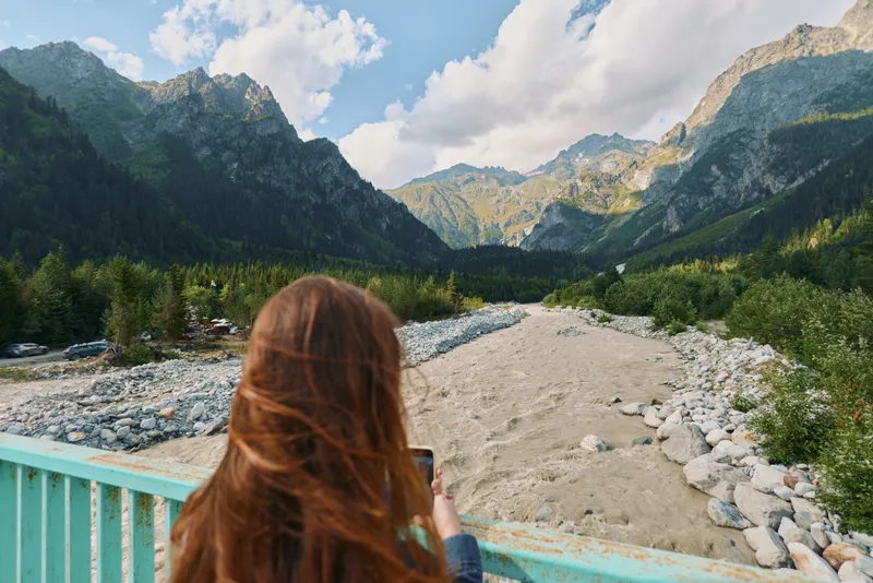 A traveler stands on a bridge, taking a photo of the surrounding mountains. The back view emphasizes focus and immersion in nature, capturing a moment of outdoor exploration and serenity.