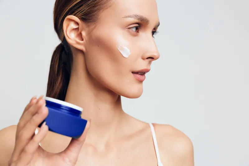 Young woman applying face cream on cheek, skincare routine, natural makeup, soft lighting, clear skin, beauty concept, white background, close up profile
