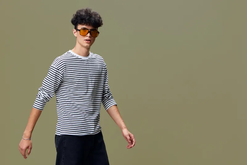 young man wearing striped shirt and sunglasses standing against plain olive background with casual style and relaxed posture