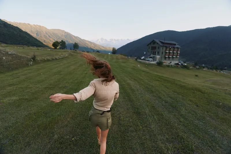 Young woman runs across a grassy hillside toward a distant chalet, wind in her hair, bright daylight, carefree energy, open space and expansive mountains.