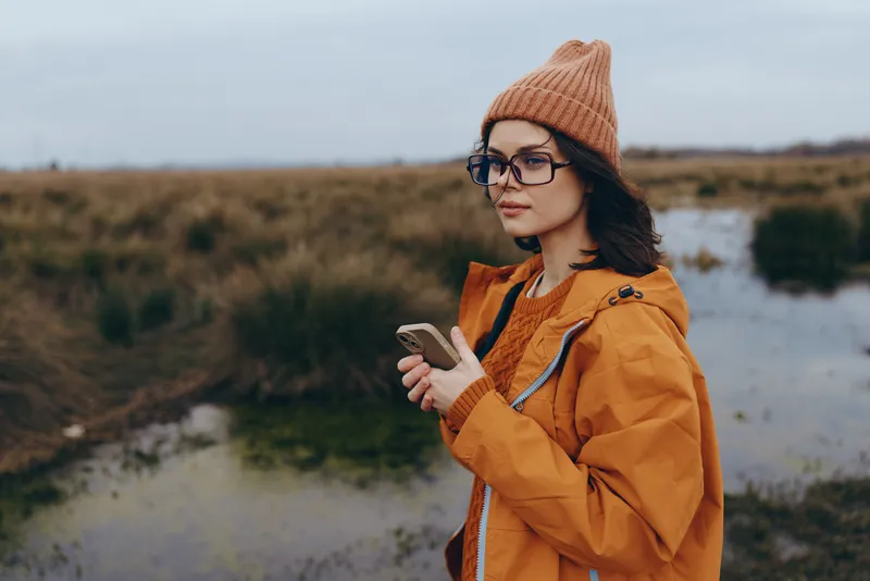 Woman lifestyle outdoors in autumn, wearing orange jacket and knitted hat, glasses on her face, holding smartphone, looking aside in scenic landscape with cloudy sky, calm mood, slow walk