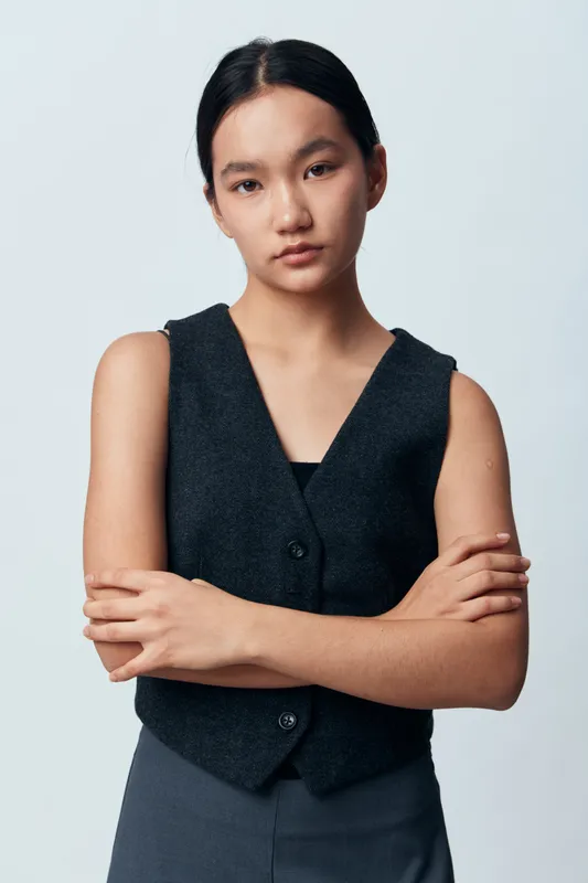 Young woman with straight black hair wearing dark vest and pants standing with arms crossed on light background in studio. Serious expression, isolated, casual fashion and confident pose.