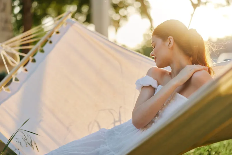 Serene young woman relaxing in a hammock at sunset, wearing a flowing white dress, with a calm expression, surrounded by greenery, evoking tranquility and summer vibes