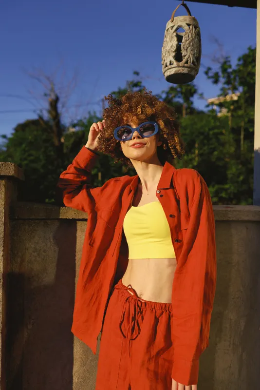 Smiling woman with curly hair wearing a yellow crop top and orange jacket outdoors, sunglasses on a sunny day. Casual lifestyle fashion, confidence, and playful style