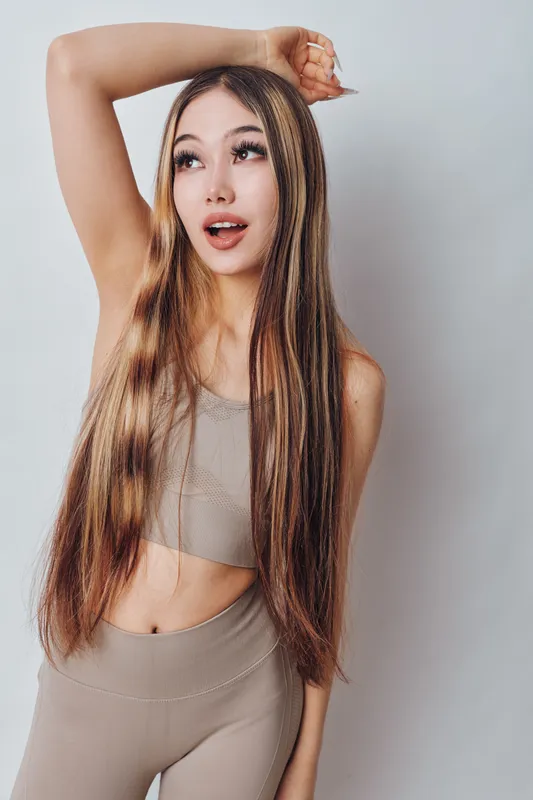 Young woman with long hair and braids wearing beige crop top and leggings posing with arm raised against light background for fashion and beauty concept.