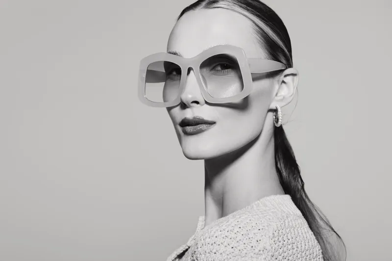 Stylish white woman with pale skin wearing large square sunglasses looks confident. Close-up portrait in black and white studio setting. Concept of fashion and elegance.