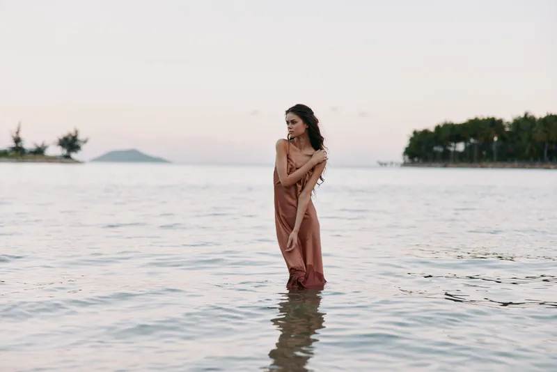 A graceful woman stands waist-deep in calm sea water, wearing a flowing copper dress, as soft coastal light wraps her in a serene, contemplative moment.