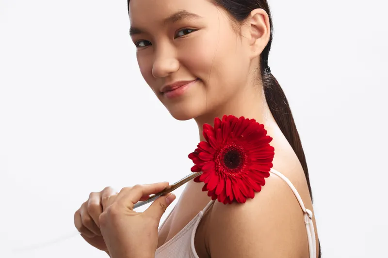 Asian woman holds a red flower near her shoulder, smiling softly against a white studio backdrop, conveying warmth, elegance, and a fresh, serene mood.