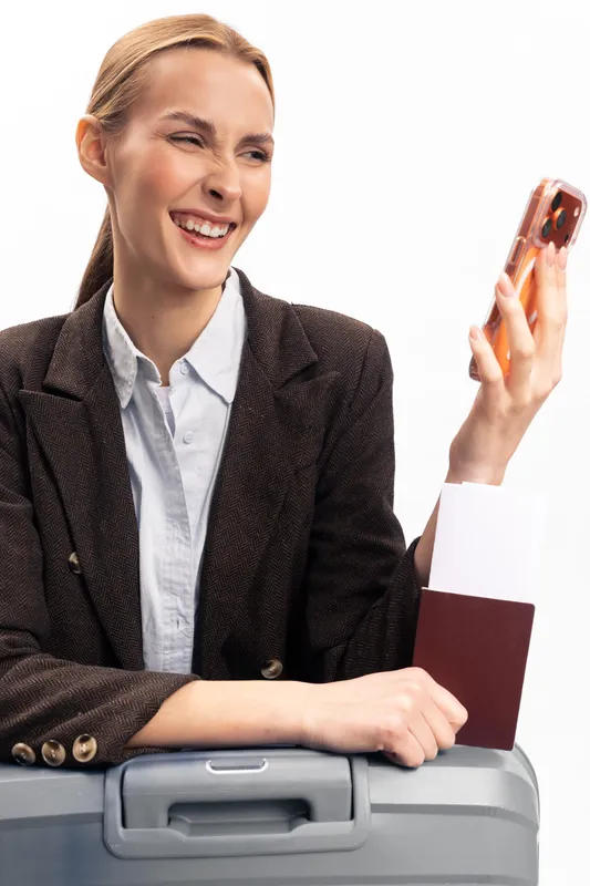 Smiling woman in blazer holds smartphone while sitting on suitcase with passport in hand. Bright white background. Concept of travel, communication, and business trip preparation.