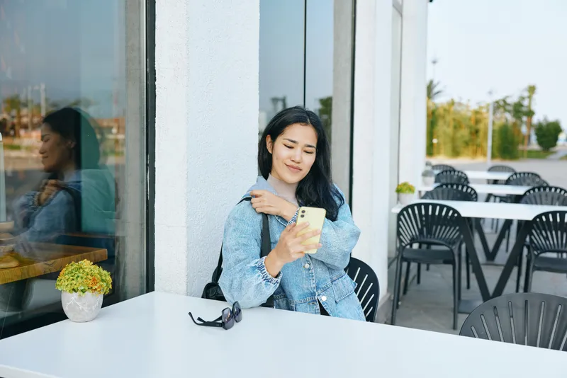 Woman seated at outdoor cafe table seen through glass, using smartphone for photo, sunglasses on table, potted plant nearby, relaxed casual mood, daytime dining, urban setting, candid moment