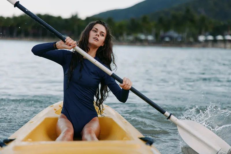 Athletic woman paddling yellow kayak on tropical river, wearing navy long-sleeve swimsuit, active lifestyle, summer adventure, water sport.