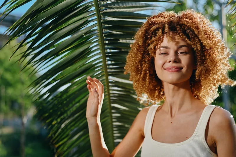 Smiling woman with curly hair enjoys sunny outdoor environment with lush green palm leaves, wearing casual white tank top, expressing happiness and relaxation.