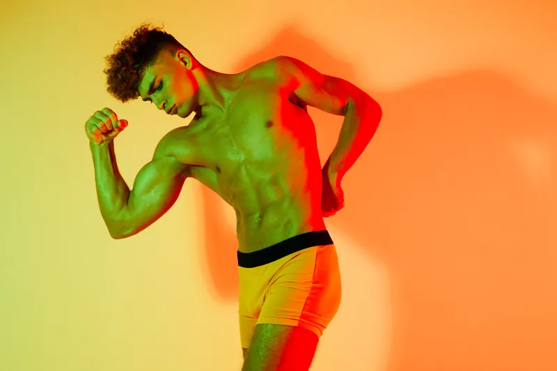 Muscular young man posing shirtless with flexed biceps and abs in colorful lighting. Athletic fit body, shorts and strong male model showing physical fitness in studio.