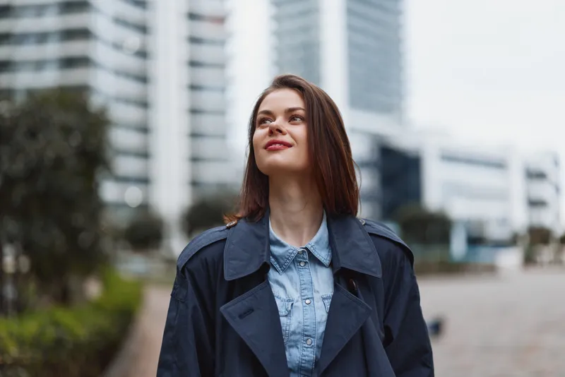 A young woman with shoulder length hair stands on an urban street, looking upward with a thoughtful smile, wearing a denim shirt under a dark trench, city buildings visible in the background.