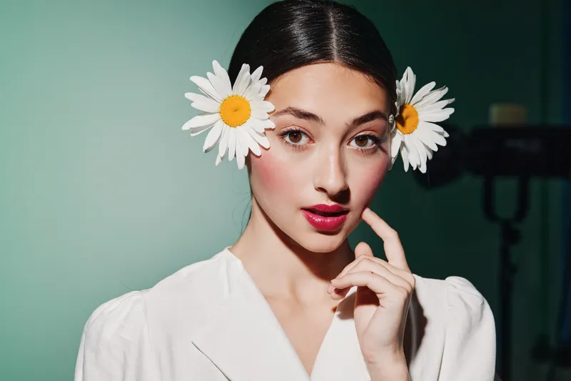Young woman in white outfit poses with two daisy flowers in her black hair against a mint background, creating a fresh beauty and fashion creative portrait concept.