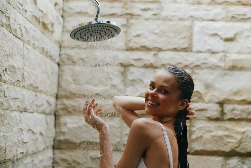 Smiling woman showering outdoors under rain showerhead with wet hair, enjoying refreshing moment against stone wall background, carefree and relaxed mood.