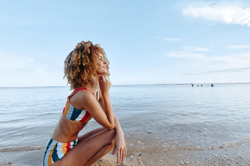 Young woman in colorful swimsuit sits on sandy beach near calm sea under blue sky, enjoying peaceful moment and warm sunlight during summer day.