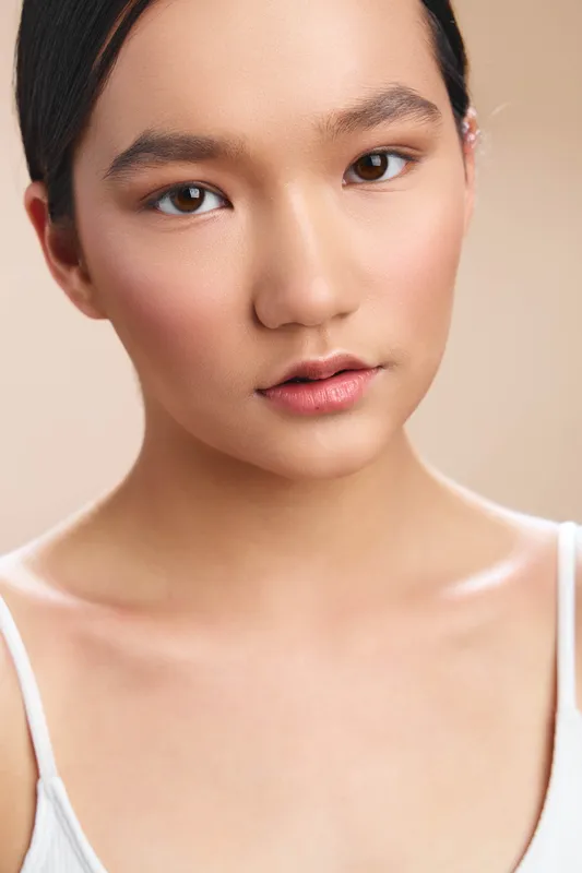 Asian woman with smooth skin gazes softly at the camera in a bright studio, conveying calm confidence and elegance. Minimal makeup and neutral tones emphasize natural beauty under gentle lighting.