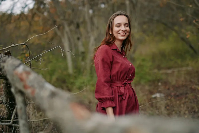 [Candid outdoor portrait of a smiling woman in a simple red dress, conveying authentic presence and natural authenticity in a quiet forest setting, focused on genuine expression and relaxed posture.]